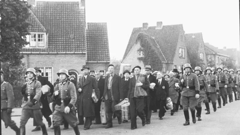 Burgers met hoeden, jassen en koffers lopen gearresteerd door een straat, ze worden begeleid door soldaten met helmen; huizen op achtergrond, sombere sfeer.