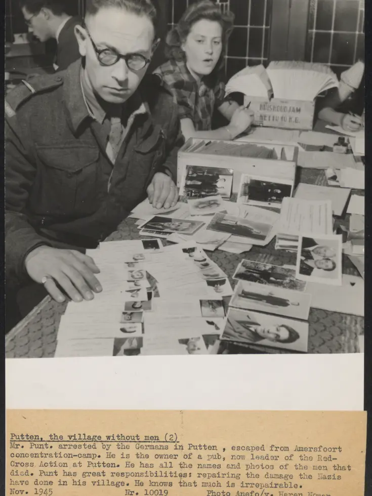 Man met bril bestudeert document aan tafel vol foto's en papieren; vrouw achter hem leest ook, geconcentreerde werksfeer.