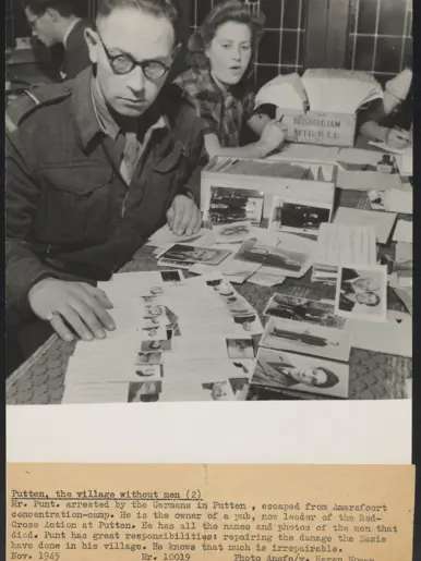 Man met bril bestudeert document aan tafel vol foto's en papieren; vrouw achter hem leest ook, geconcentreerde werksfeer.