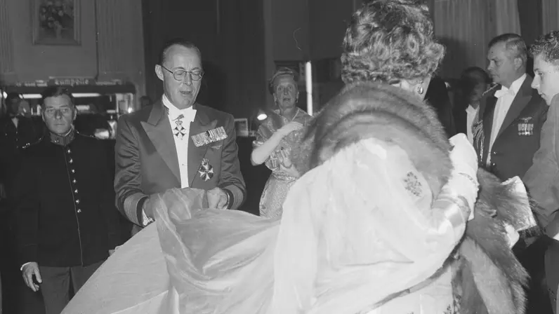 Een formeel evenement: een gedecoreerde man in pak begroet een vrouw in lange lichte jurk met bontstola, te midden van elegant geklede gasten onder een kroonluchter.