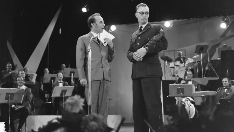 Wim Sonneveld spreekt op het toneel in een microfoon met papier in hand, naast hem kapitein Van Diepenbeek met een trofee in zijn arm. Achter hen speelt een orkest onder spotlights; het publiek kijkt als vage silhouetten.