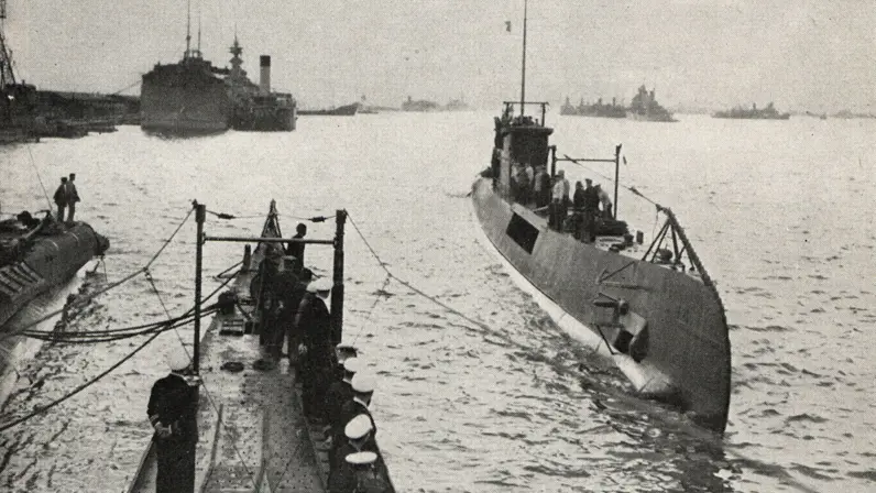 Op de voorgrond een deels ondergedoken onderzeeboot met mensen aan dek. Links zeelieden op een kade in donkere uniformen, erachter grotere marineschepen in kalm water. 