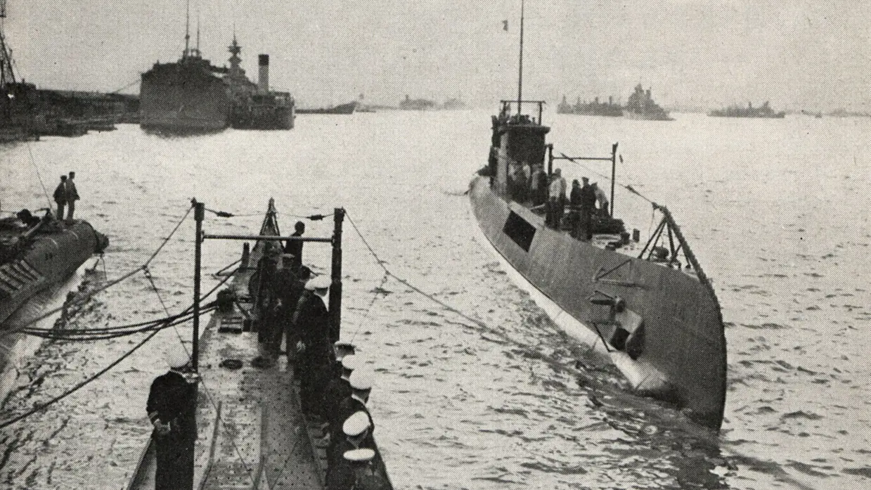 Op de voorgrond een deels ondergedoken onderzeeboot met mensen aan dek. Links zeelieden op een kade in donkere uniformen, erachter grotere marineschepen in kalm water. 