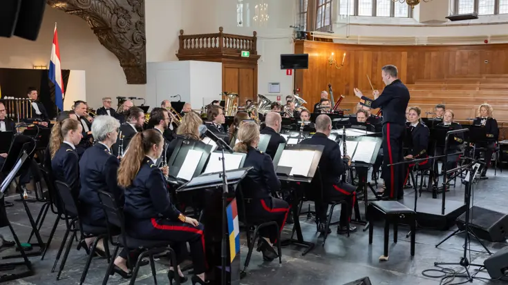 Dirigent staat voor militair orkest dat zittend speelt.