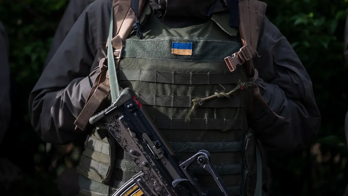 Close-up van de torso van een militair. Hij draagt een scherfwerend vest, een geweer en een Oekraiëns vlaggetje op zijn uniform.