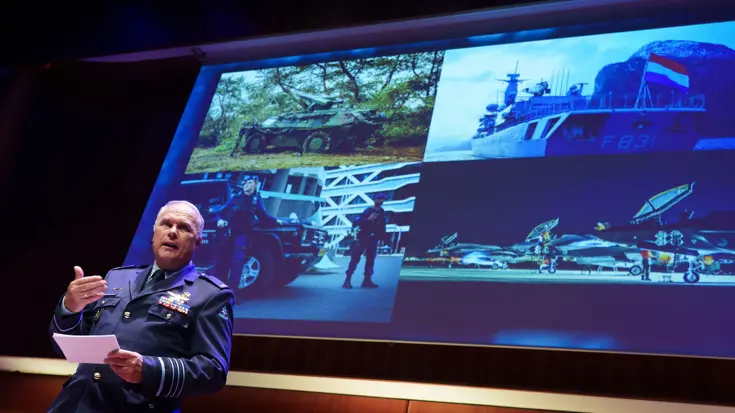 Eichelsheim staat op het podium voor een groot scherm en spreekt de zaal toe. 