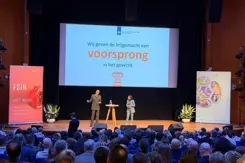 Luitenant-generaal Jan-Willem Maas staat op een podium met achter hem een groot scherm. Op de voorgrond van de foto achterhoofden van mensen in de zaal.