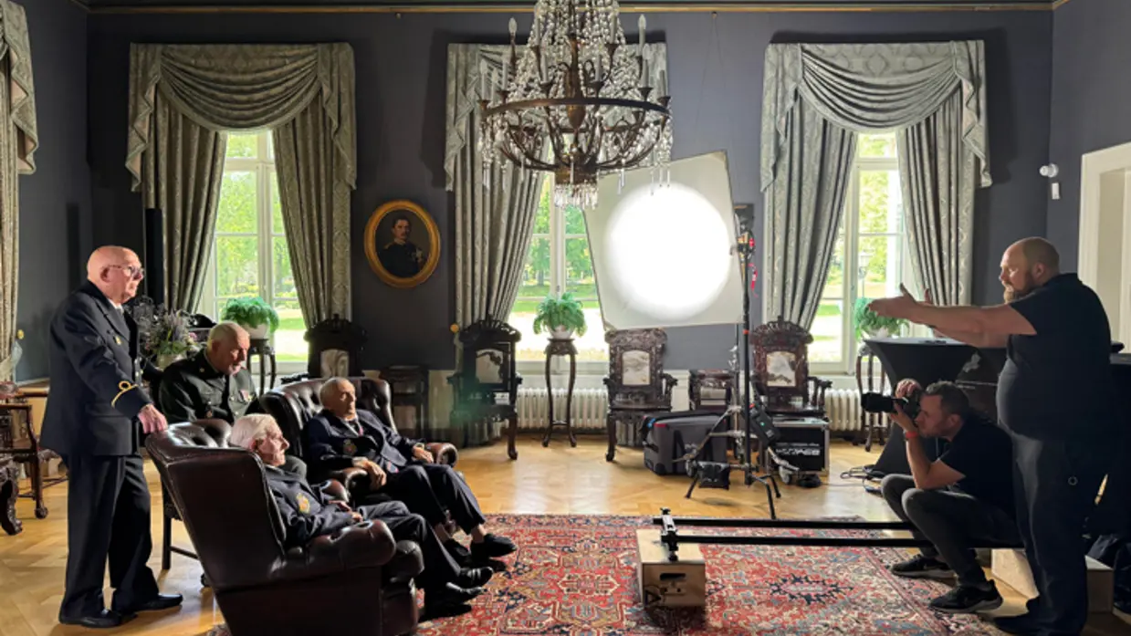 Regisseur Joas Burggraaf staat tegenover een groep oude veteranen. Zij staan in beeld van een camera onder een grote lamp in de commandantenkamer van Bronbeek.&nbsp;