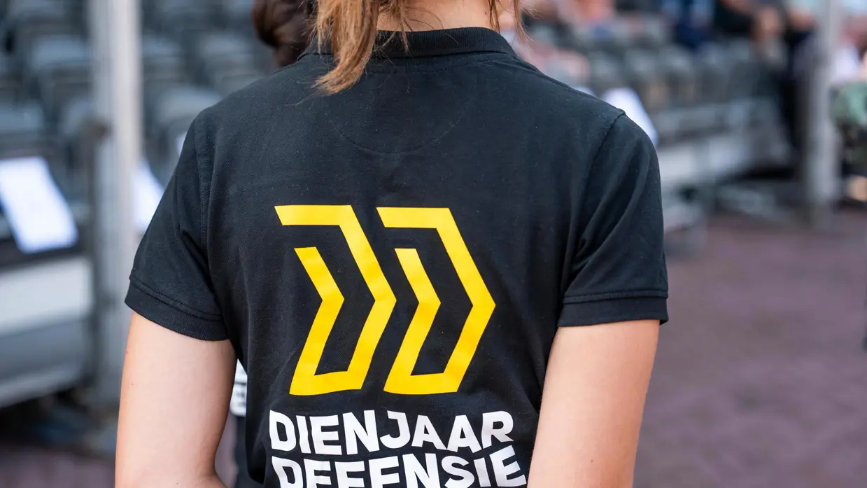 Rug van vrouw die polo draagt met daarop geel logo en tekst Dienjaar Defensie.