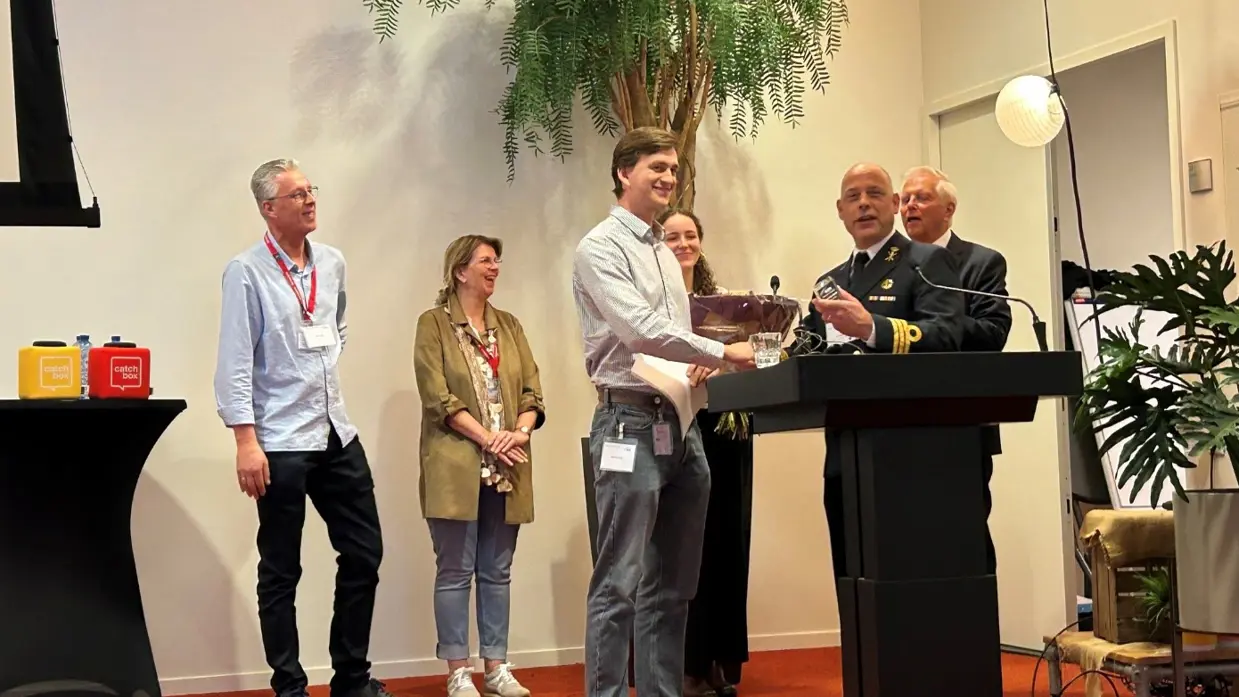 Student Jasper en juryvoorzitter Ivo schudden elkaar de hand op een podium. Op de achtergrond staan familieleden van de winnaar.