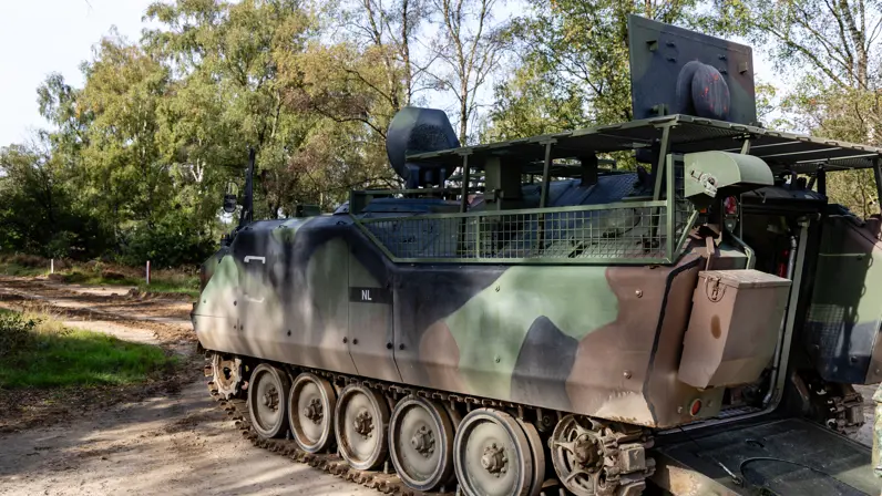 Een gecamoufleerd voertuig op rupsbanden staat op de weg met bosschages op de achtergrond. 