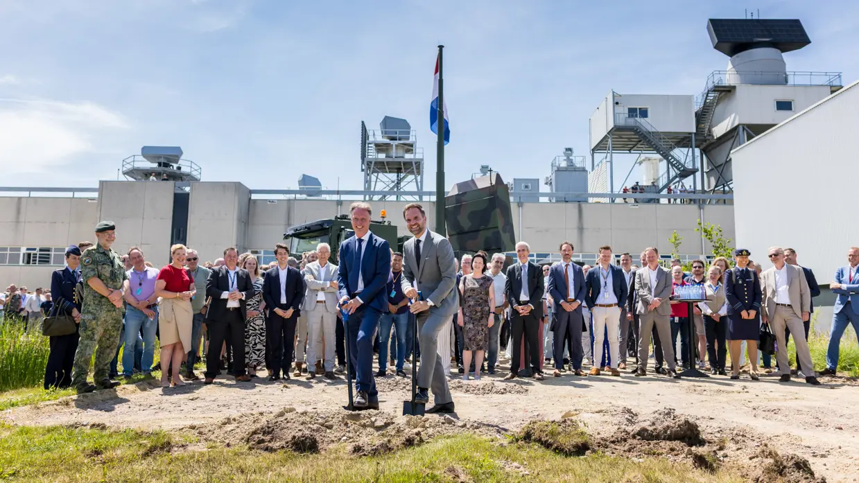 Twee mannen in net pak steken een schop in de grond. Daarachter een toekijkende mensenmenigte.