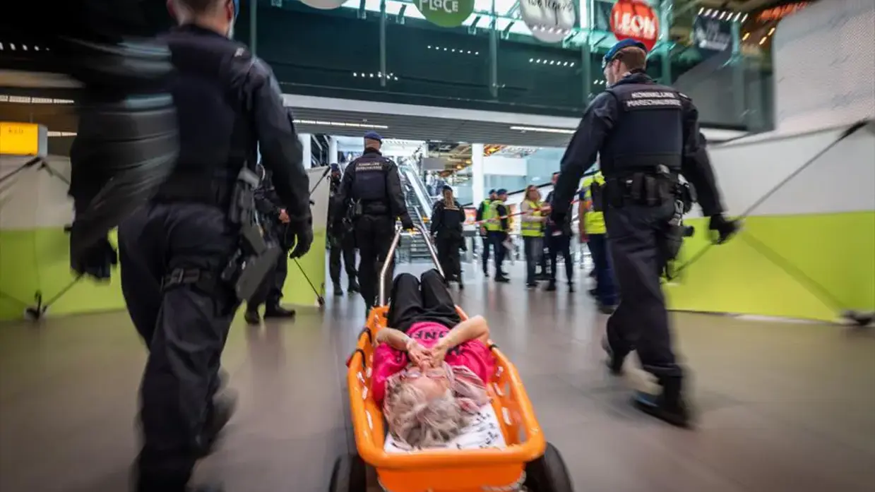 Demonstranten liggen op Schiphol met een scherm om hen heen.