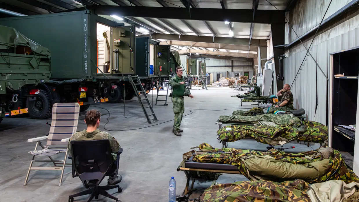 Opgerolde slaapzakken liggen op veldbedden in een militaire hal. Groene legervoertuigen en open container links. Soldaten zitten op stoelen te rusten in deze ordelijke, rustige ruimte.
