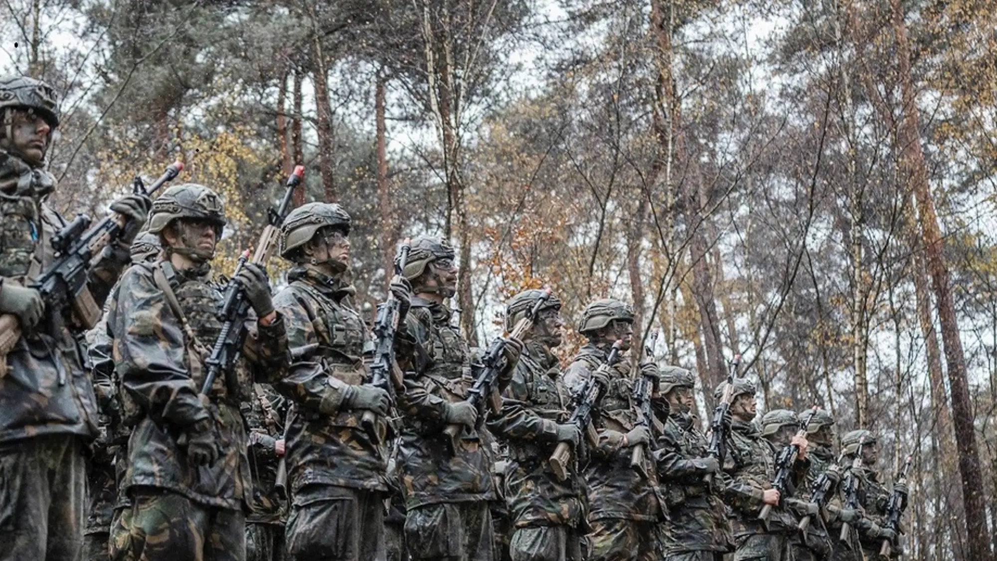 Groep militairen met camouflagekleding en -schmink houden wapens voor de borst terwijl ze in een bos in rijen staan opgesteld.