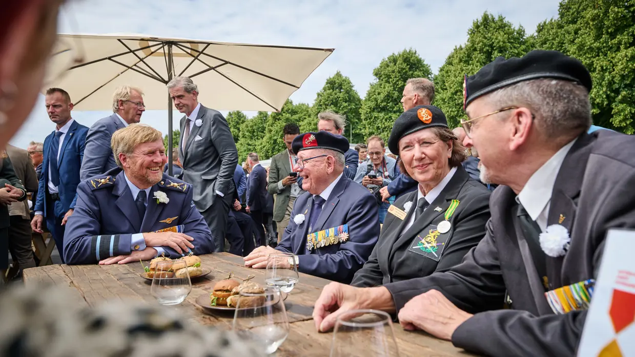 Een lachende koning Willem-Alexander aan een picknicktafel in gesprek met een veteraan.