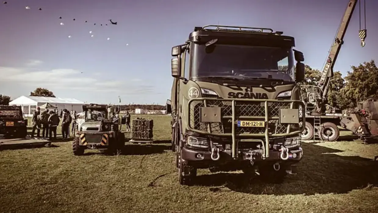 Een militaire truck met op de achtergrond vliegtuigen en parachutisten.