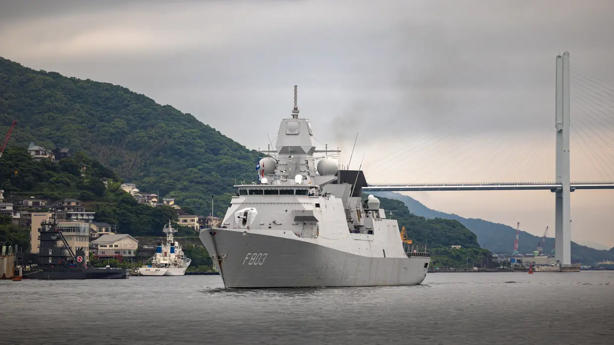 Een varend, grijs marineschip. Op de achtergrond een brug en groene heuvels.