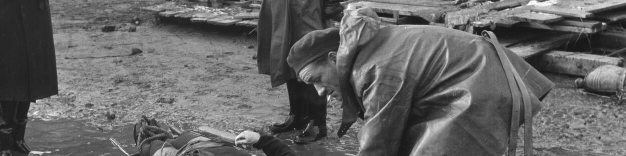 Zwart-witfoto. Man probeert in een plas water een brancard met een slachtoffer op te tillen na de watersnoodramp van 1953.