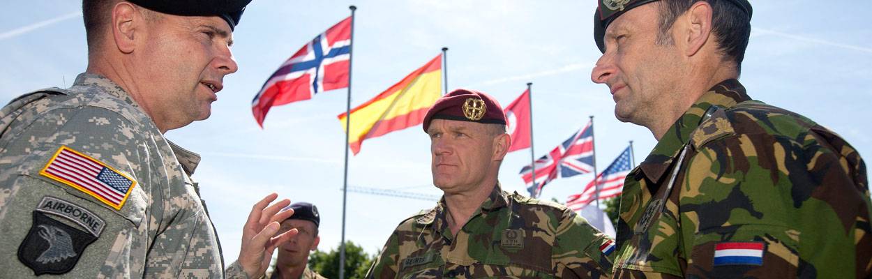 Nederlandse en Amerikaanse militairen praten met op de achtergrond een rij met internationale vlaggen.