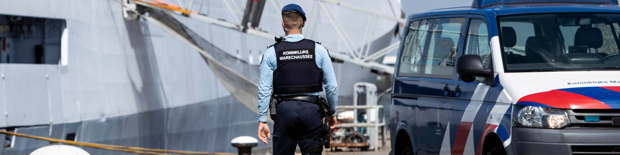 Een marechaussee loopt op een kade. Links ligt een schip, rechts staat een wagen van de Koninklijke Marechaussee.