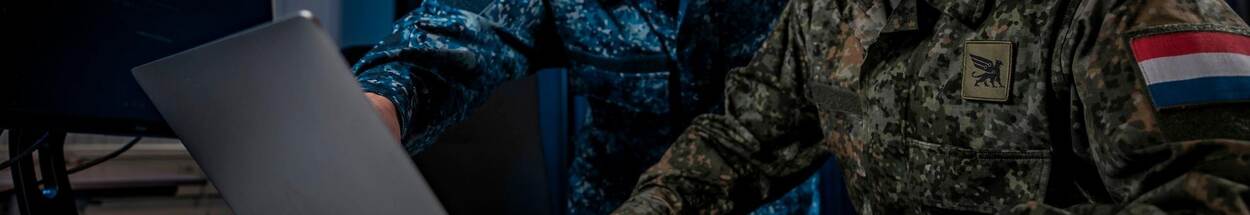 Militair in groen camouflagepak en militair in blauw camouflagepak achter beeldscherm.