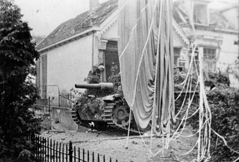 Zwart-witfoto: op de voorgrond een bevoorradingsparachute van de geallieerden. Op de achtergrond een tank voor een gehavend woonhuis.