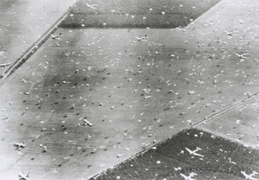 Zwart-wit luchtfoto van de geallieerde dropzone bij Arnhem. Te zien is een veld waarboven laag vliegtuigen vliegen die talloze parachutisten en voorraden droppen, maar ook al gelande zweefvliegtuigen en parachutisten.