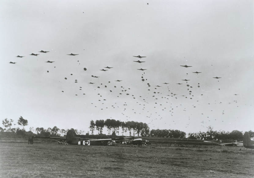 Historische zwart-wit foto vanaf de geallieerde dropzone. Op de voorgrond liggen al gelande zweefvliegtuigen in een grasveld met een paard. In de lucht op de achtergrond zijn 10-tallen geallieerde vliegtuigen te zien die talloze parachutisten droppen.