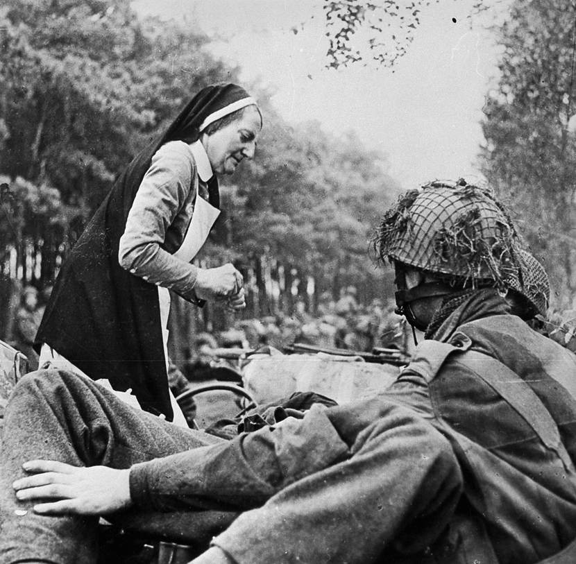 Zwart-witfoto: een Nederlandse Rooms-Katholieke non ontfermt zich over een gewonde geallieerde militair op een voertuig, onder het toeziend oog van 2 andere geallieerde militairen.