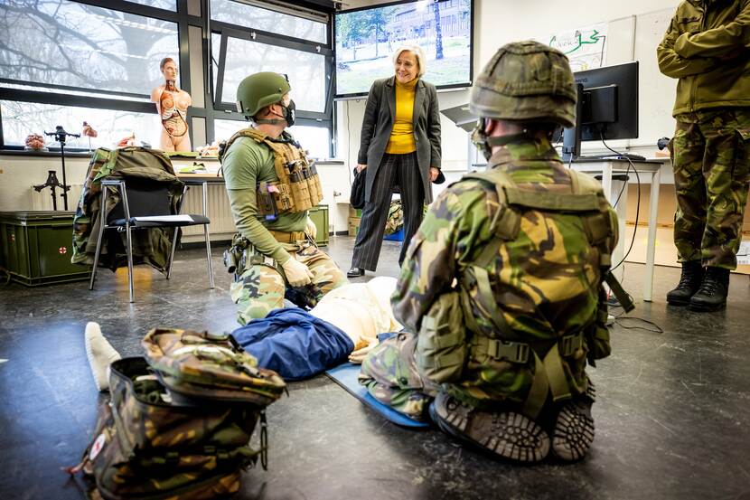 Minister Bijleveld in een opleidingsruimte voor bedrijfshulpverlening. 2 militairen zitten op de grond aan weerszijden van een beademingspop.
