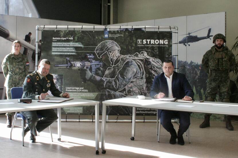 Militair en burger zitten aan een tafel voor het ondertekenen van een contract. Op de achtergrond een poster van STRONG.