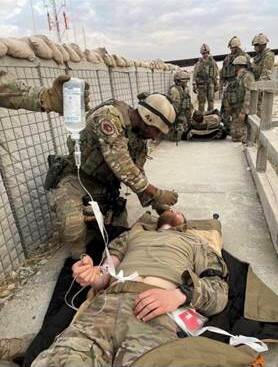 Een gewonde militair met een infuus op een brandcard. Collega-militair verzorgt de gewonde.