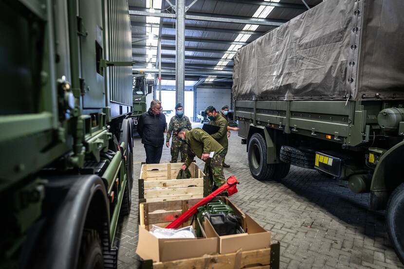 Militairen kijken tussen spullen in kisten, die tussen 2 militaire voertuigen staan.