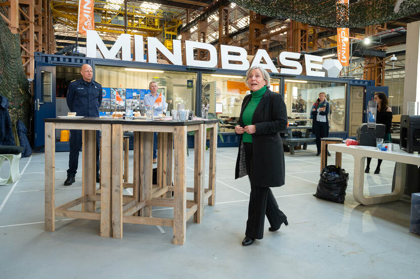 De minister loopt naar een tafel, op de achtergrond in grote letters 'Mindbase'.