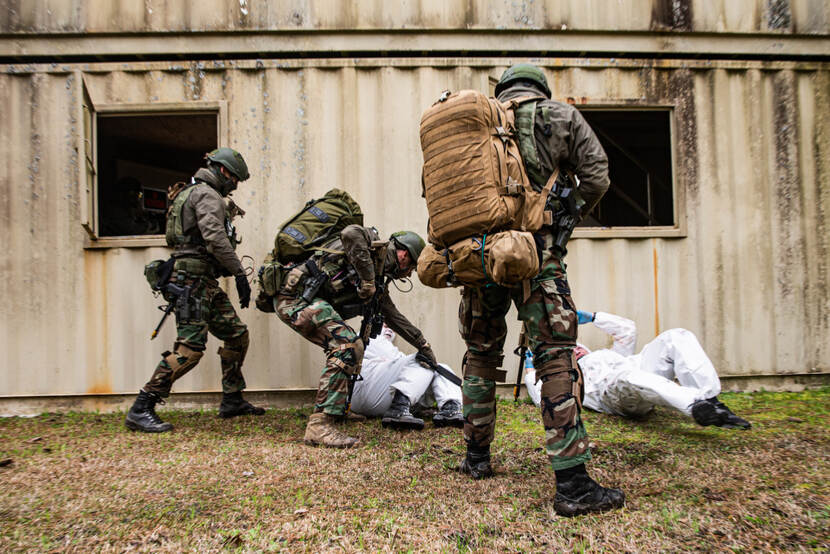 3 bewapende militairen houden 2 mannen in witte pakken liggend op de grond.