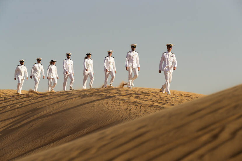 Marinepersoneel in witte uniformen lopen over een zandduin in de woestijn.