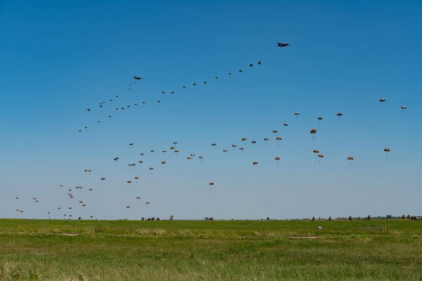 Tientallen parachutisten, gedropt uit vliegtuigen, zweven boven een grasveld.