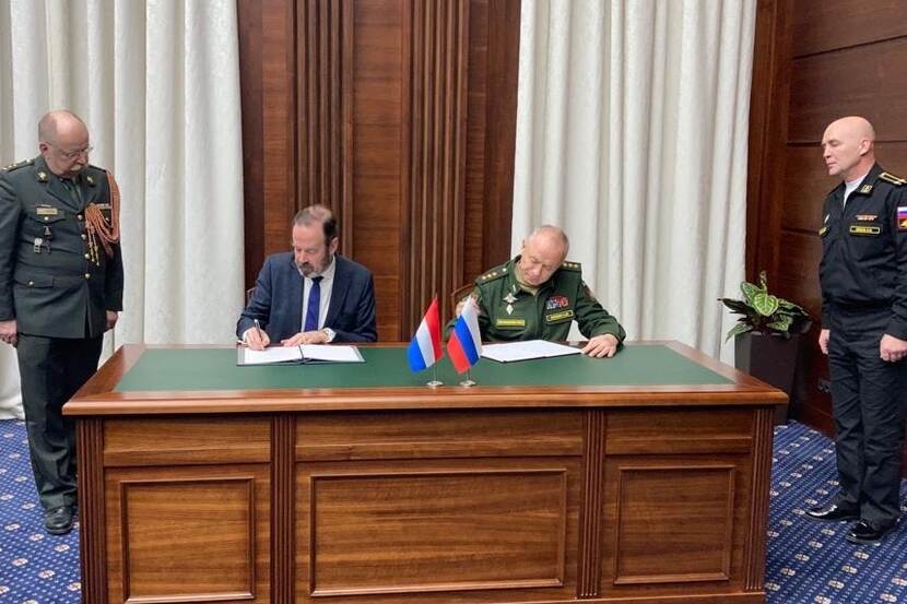 2 heren zitten achter een zwaar houten bureau en tekenen de gewijzigde overeenkomst, 2 militairen kijken toe.