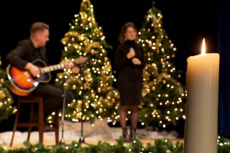 Screenshot kerstviering Defensie 2021. Kaars met op achtergrond gitarist en zangeres.