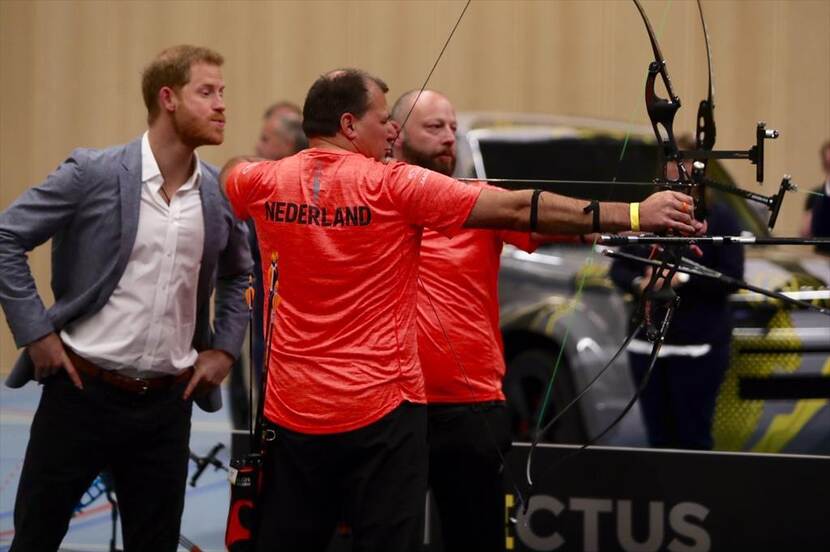 Prins Harry kijkt mee over de schouders van 2 Nederlandse boogschieters.