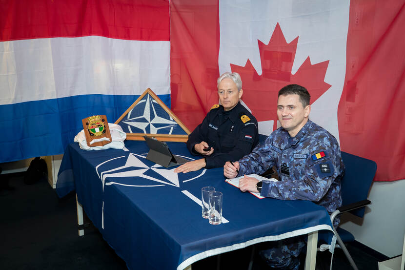 2 militairen zitten aan een tafel met daar overheen een NAVO-vlag. Erachter hangen de vlaggen van Nederland en Canada.
