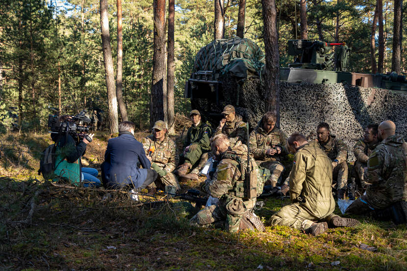 Groep militairen, zittend in een bos, met cameraman en interviewer.