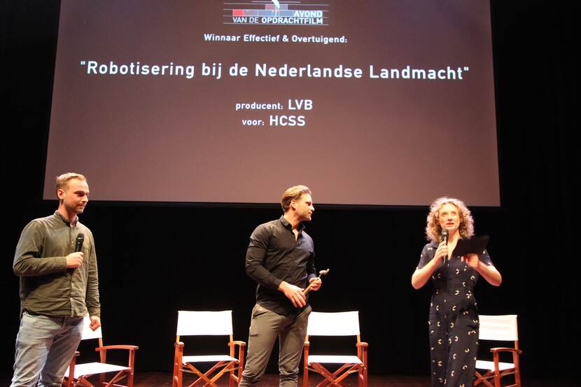 3 mensen op een podium, met op de achtergrond een beeld met de tekst: Winnaar effectief en overtuigend "Robotisering bij de Nederlandse Landmacht".