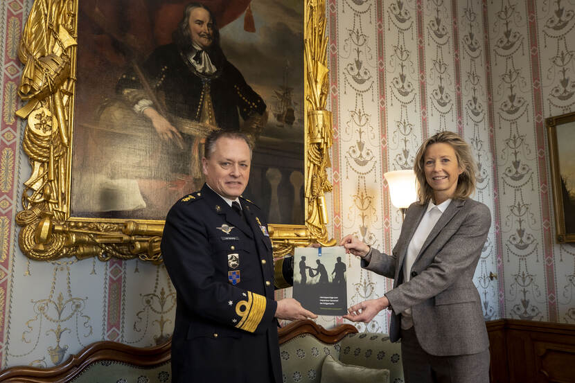 Minister Kajsa Ollongren krijgt van luitenant-generaal Frank van Sprang het IGK-jaarrapport, in een werkkamer met een historisch schilderij op de achtergrond.