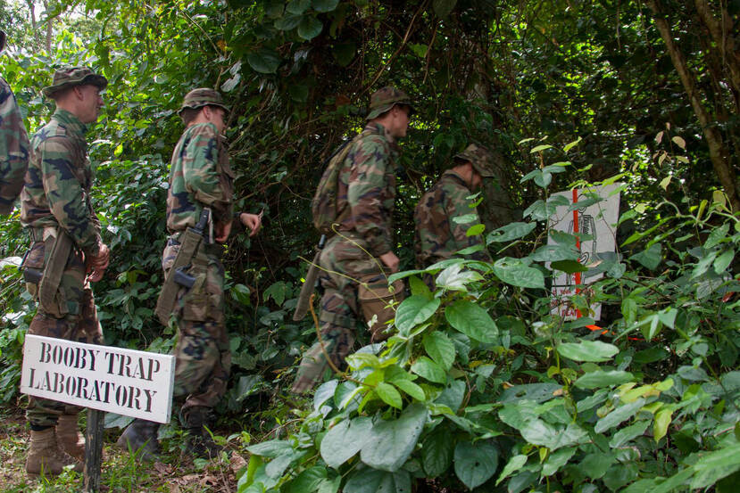 Militairen in een bos lopen langs een bordje met de tekst booby trap laboraty.