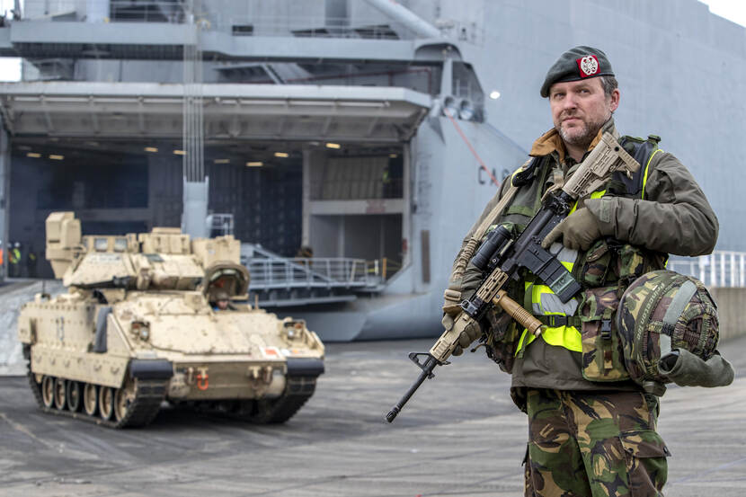 Nederlandse militair met wapen staat voor een Amerikaanse tank, met op de achtergrond een marineschip.
