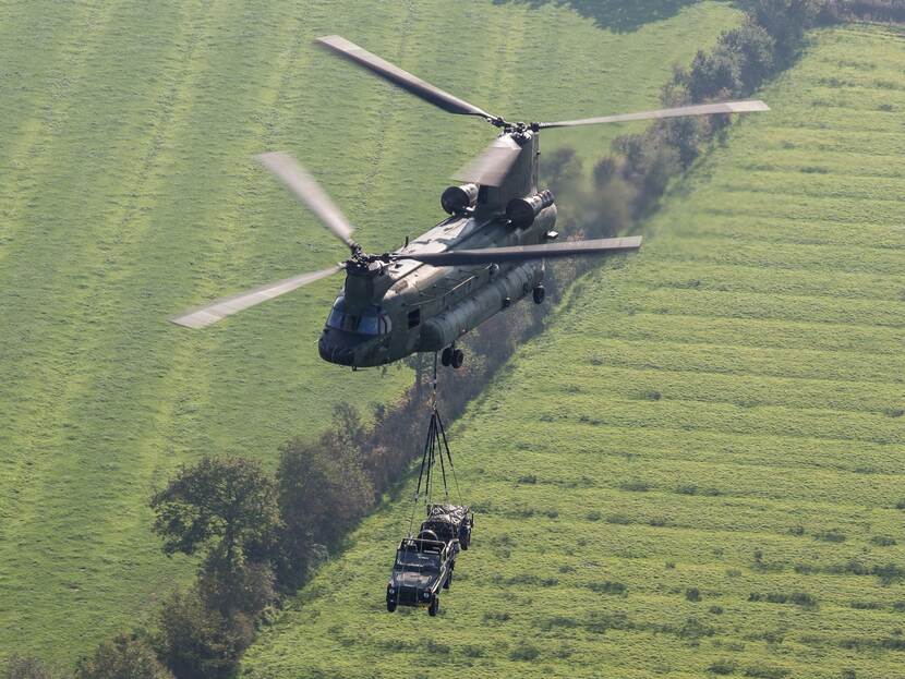 Bovenaanzicht van vliegende Chinook, eronder hangt een militair voertuig.