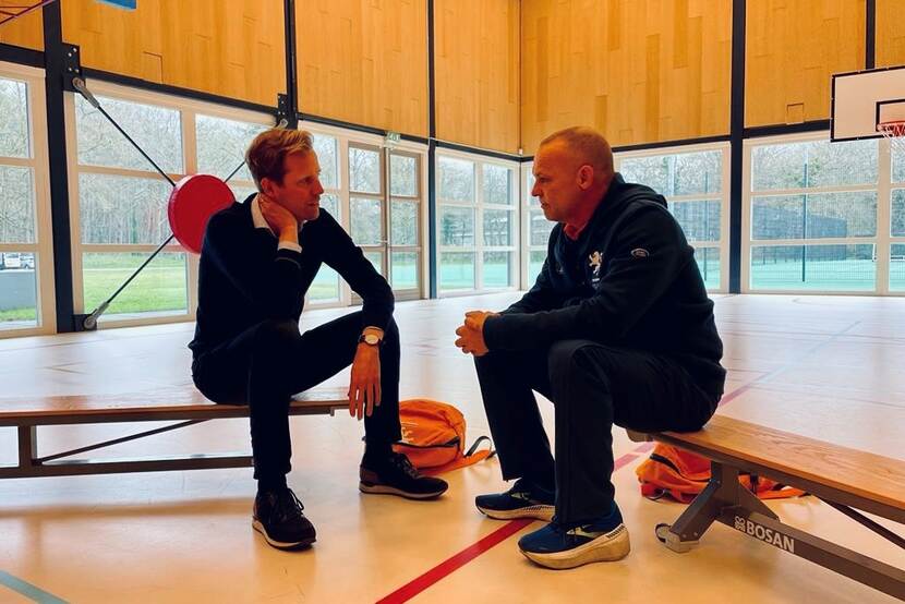 Staatssecretaris Christophe van der Maat zittend op een bank in een sportzaal in gesprek met een Invictus-sporter.