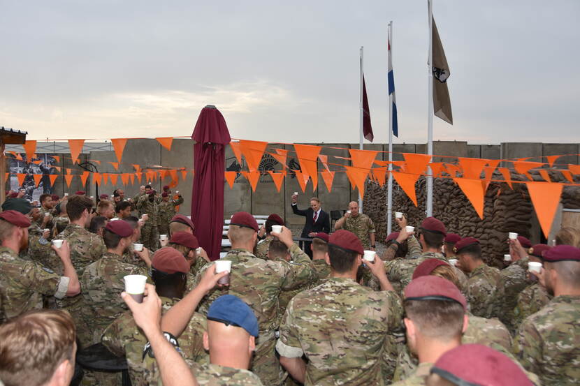 Groep militairen toost met plastic bekertjes. Oranje vlaggetjes boven hen.